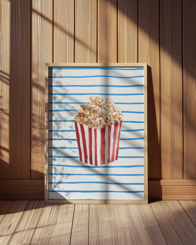 Playful popcorn art print featuring a red and white striped popcorn box against a white background with blue horizontal lines. Displayed in a light wooden frame leaning against a wooden wall, illuminated by warm natural light. Perfect kitchen or movie room wall art with a retro touch.